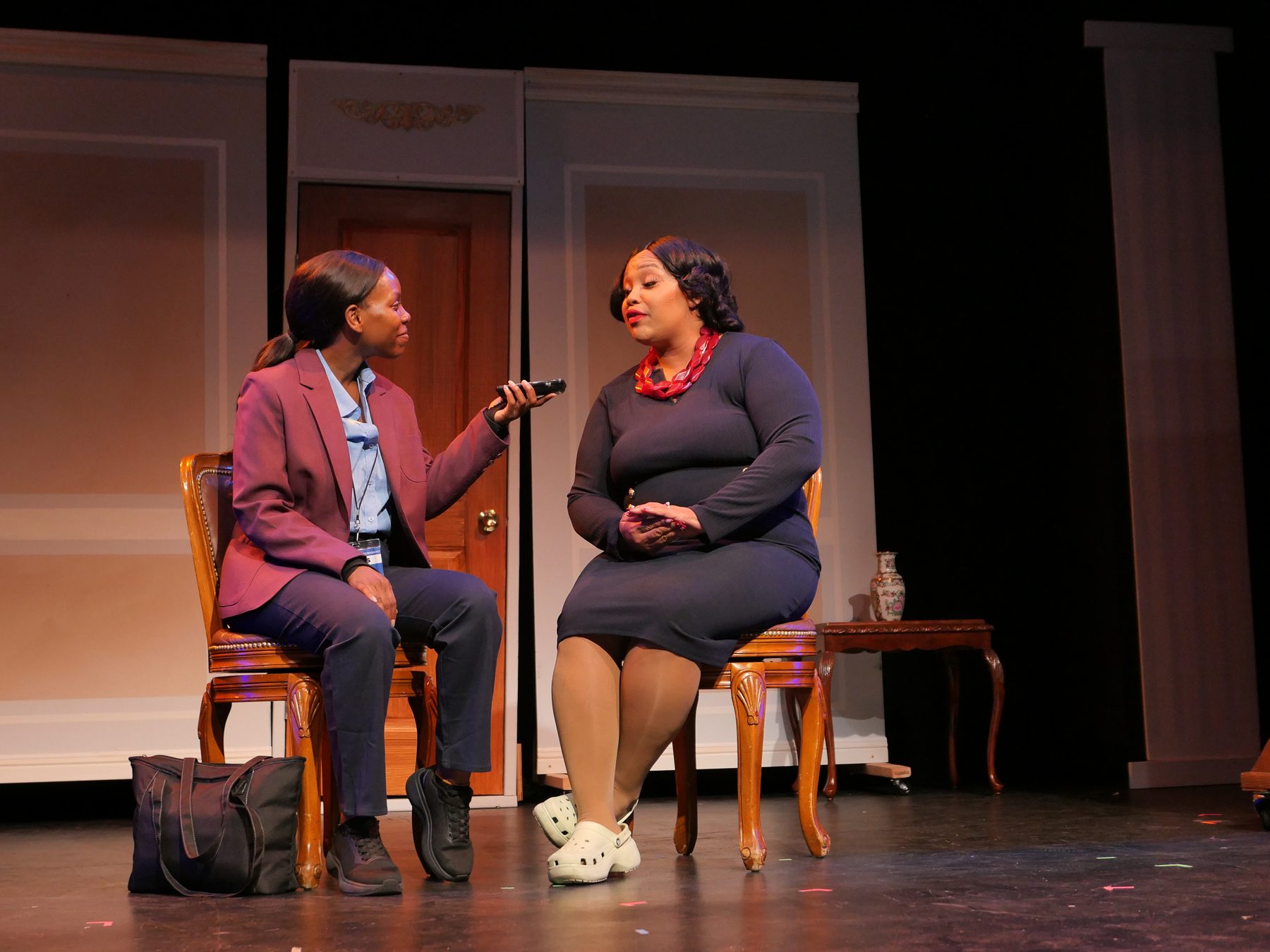 A reporter in a burgundy blazer holds out a recorder to a woman in a navy dress seated beside her.